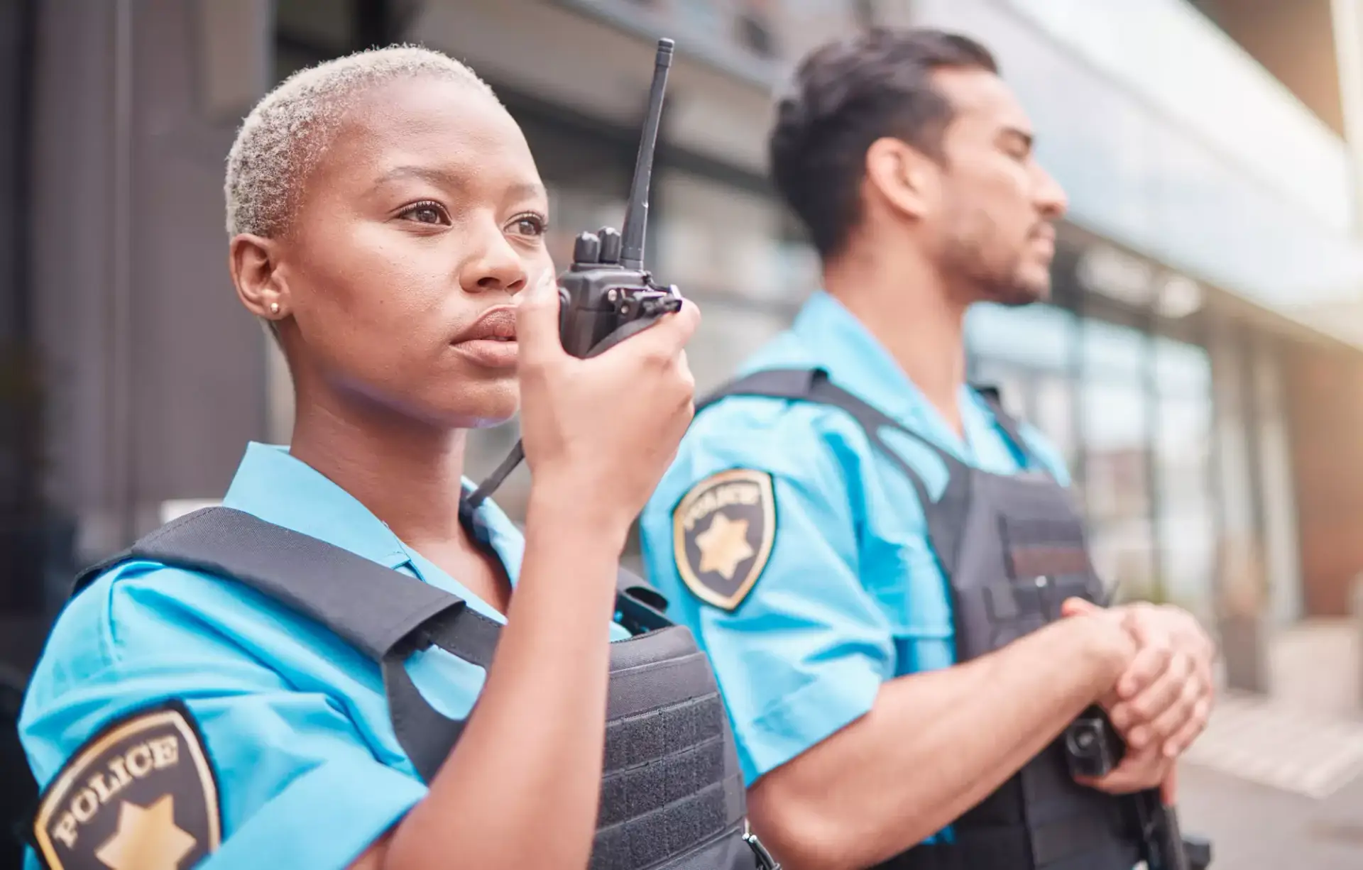 Two police officers in light blue uniforms and tactical vests, with one officer using a handheld radio.