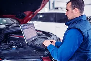 A mechanic in a blue uniform using a diagnostic laptop to analyze a car engine in an auto repair shop.